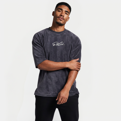 Sportliches Herrenmodel im Elite Club Oversized Cord T-Shirt Anthrazit mit weißem Brustlogo, Arm locker überkreuzt, moderne Streetwear Studioaufnahme