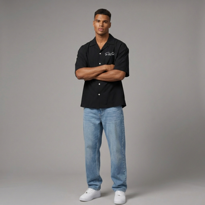 Selbstbewusstes männliches Model mit verschränkten Armen trägt schwarzes Elite Club Kurzarmhemd, kombiniert mit Jeans und weißen Sneakern, minimalistisches Studio Setting.