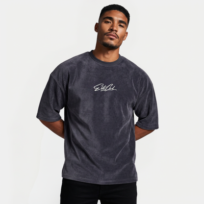 Männliches Model mit geneigtem Kopf trägt anthrazitfarbenes Elite Club Cord T-Shirt mit Logo Print, Hände hinter dem Rücken, Studioaufnahme