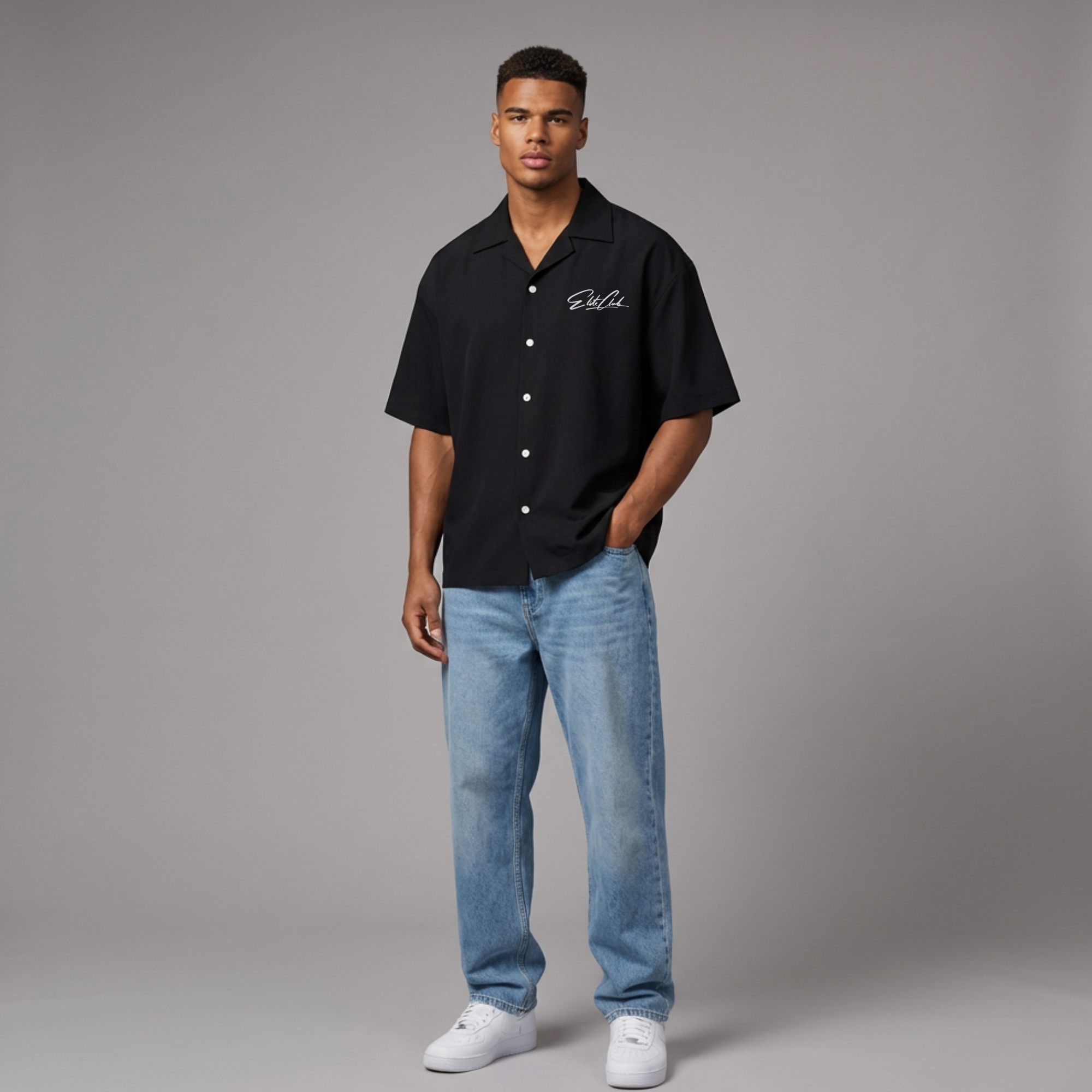 Model in schwarzem Elite Club Kurzarm Button-Up Hemd mit Brustlogo, helle Jeans und weiße Sneaker, lässige Pose mit Hand in der Hosentasche.