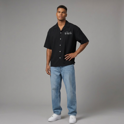 Männliches Model trägt schwarzes Elite Club Kurzarmhemd mit Logo-Stickerei, kombiniert mit hellblauer Jeans und weißen Sneakern vor neutralem Studiohintergrund.