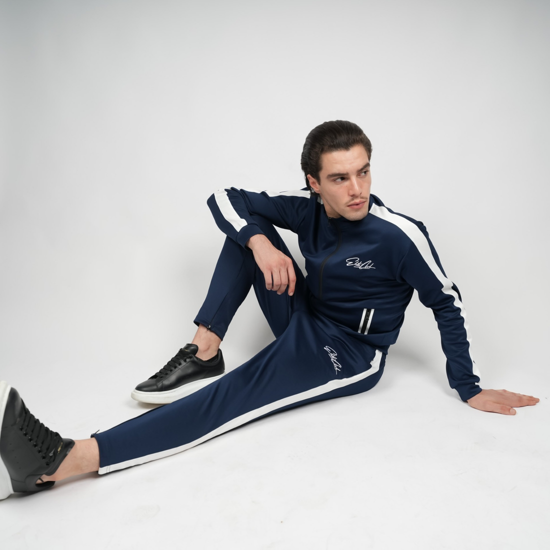 Herren Elite Club Performance Trainingsanzug in Navy Blau mit weißen Streifen, sportliche Slim-Fit-Passform, sitzende Pose im Studio